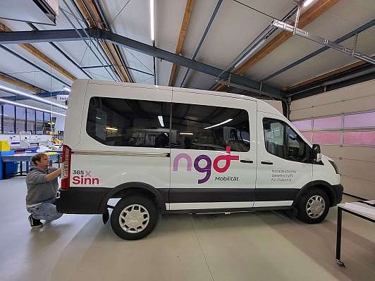 Ein Kleinbus in einer Werkstatthalle wird von einem Mann mit neuem Logo beklebt